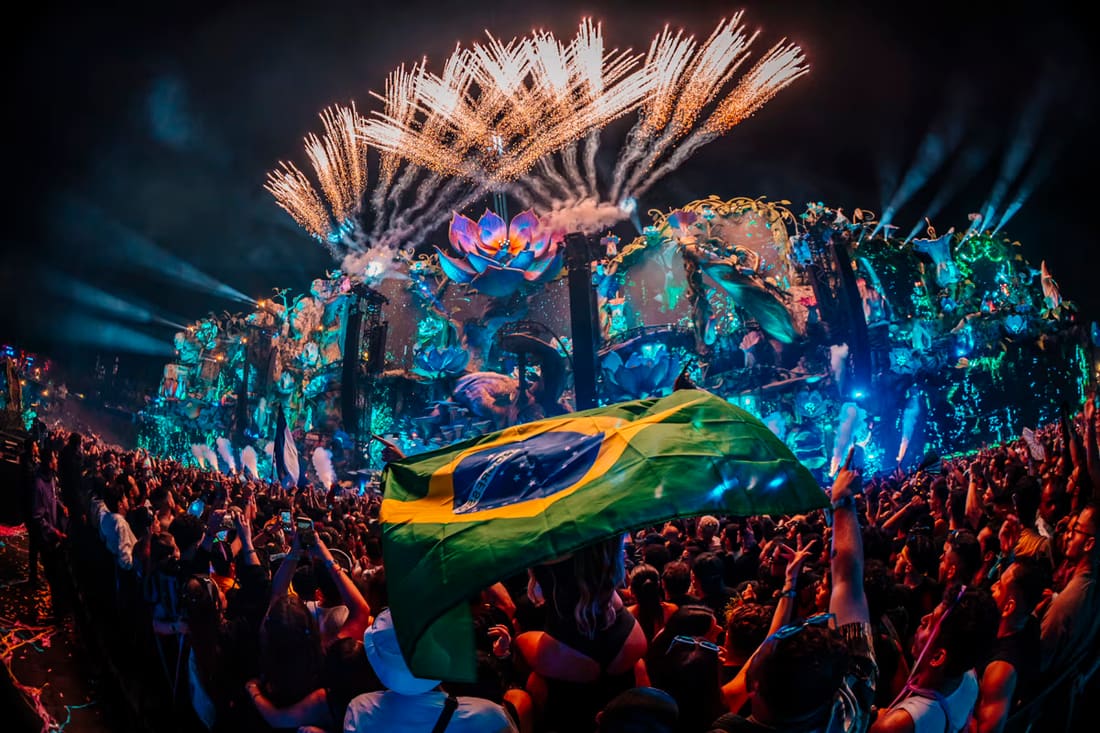 A bandeira do Brasil erguida no meio do público, com fogos sobre o Mainstage LIFE do Tomorrowland Brasil 2025, à noite.