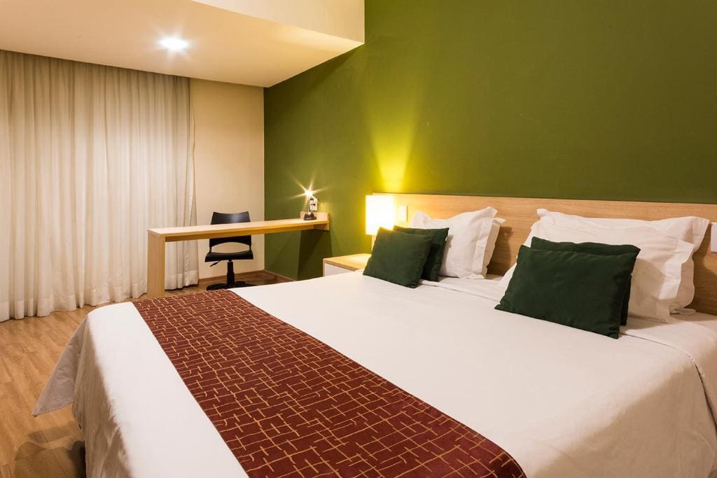 Apartamento duplo casal do Hotel Never Ends: cama de casal com almofadas verdes, mesa de trabalho e cortina até o chão.